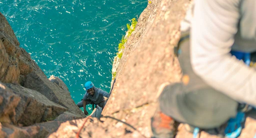 A person wearing safety gear is secured by ropes as they rock climb. They are looking up at the camera and there is blue water below them.